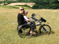 Kristina wearing sunglasses on a beautiful sunny day, sat in a wheelchair with Bike adaption in the middle of green fields with a young child sat on her lap.