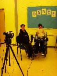 two young people, one is a wheelchair user, are sat smiling facing a camera on a tripod with the word DANCE on the wall behind them