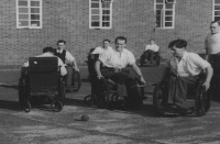 Black and White photograph of six young men, smartly dressed in white shirts and dark trousers playing wheelchair polo